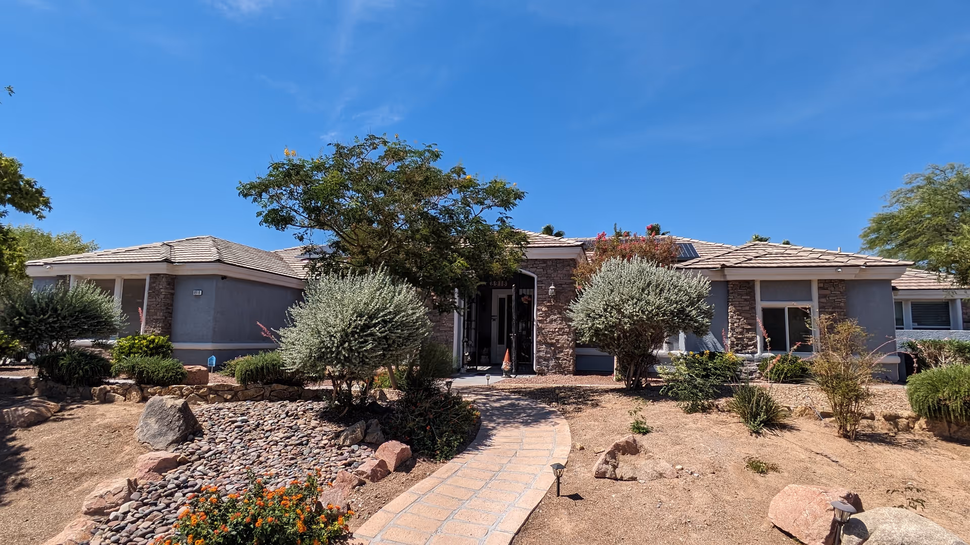 Front exterior view of a single-story senior home building with a tiled roof, stone accents around the entrance, and a paved walkway leading to the front door. The landscape includes desert-style plants, bushes, rocks, and a clear blue sky overhead.