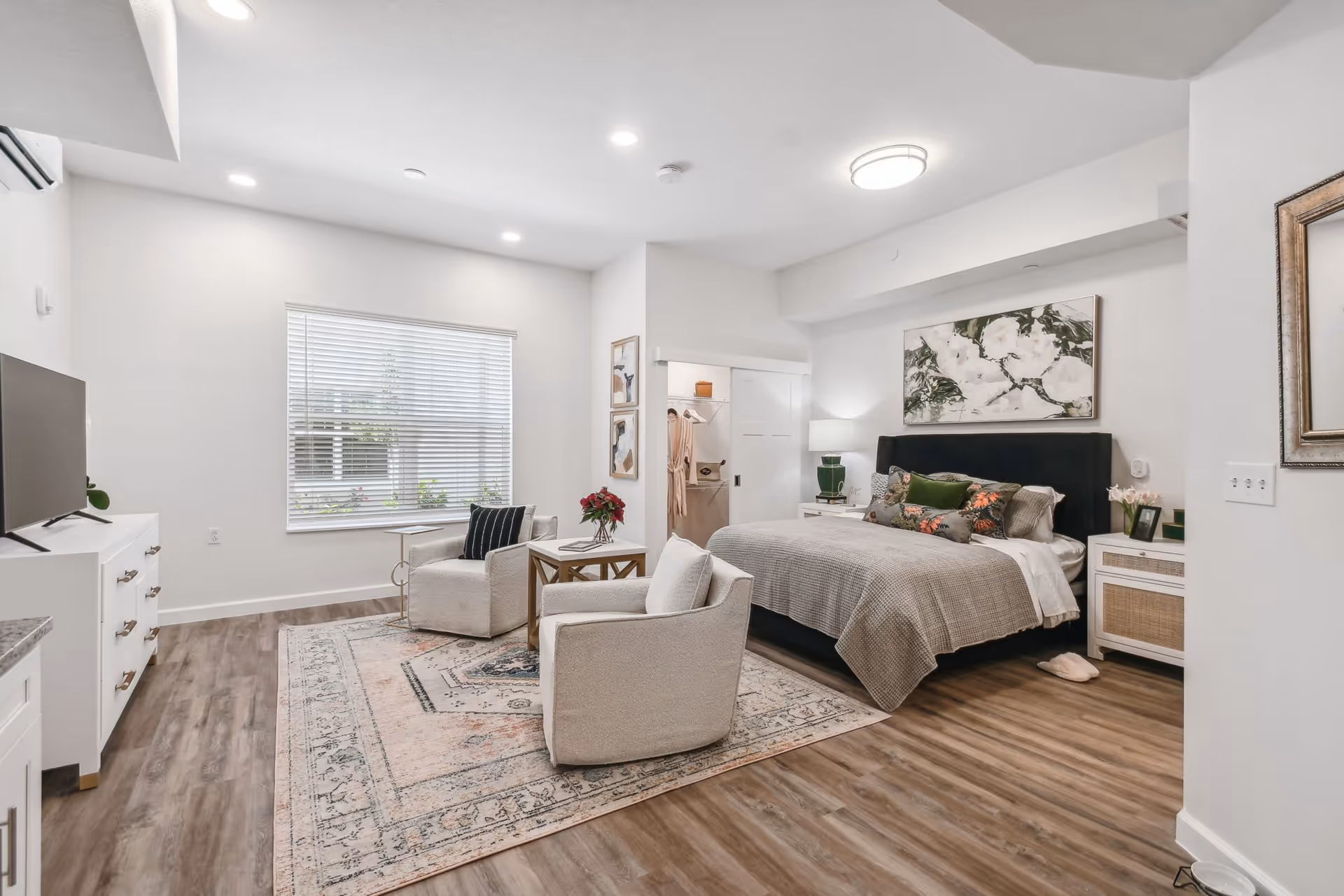 A bright and modern bedroom with a large window covered by blinds, two beige armchairs with a small wooden table between them on a patterned area rug, a bed with a dark upholstered headboard and floral pillows, nightstands with lamps, and a closet with sliding doors. The room has wood flooring and white walls with minimal decor.