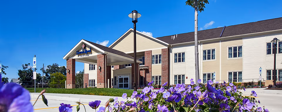 Exterior view of Sarah Reed Senior Living facility on a sunny day with a clear blue sky. The building has a beige and brick facade with multiple windows and a covered entrance supported by brick columns. Purple flowers and greenery are visible in the foreground.