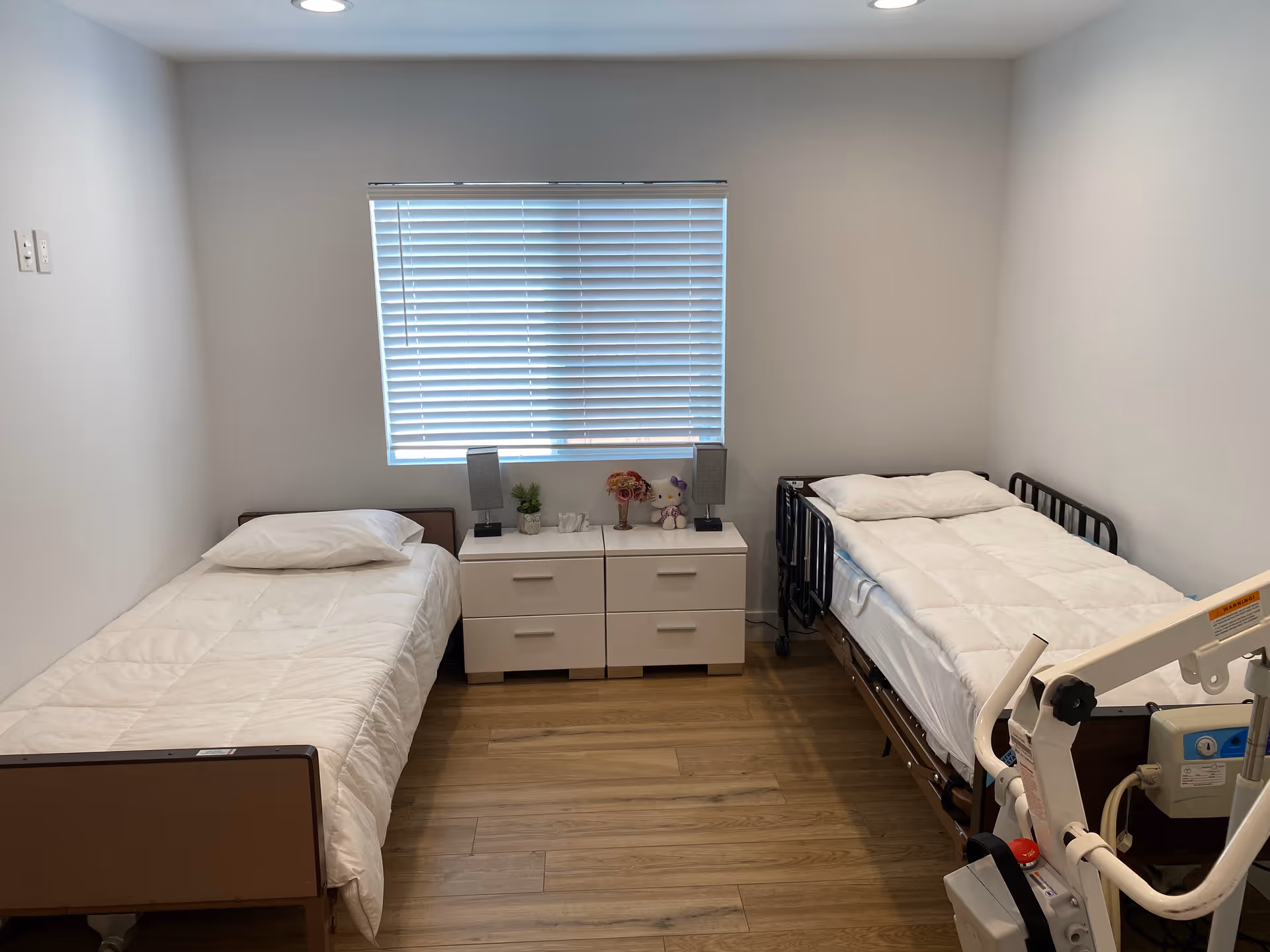 A simple bedroom with two single beds on either side of a white double-drawer nightstand. The bed on the left has a plain white comforter and pillow, while the bed on the right has a white comforter and pillow and is equipped with metal side rails. On the nightstand are two small lamps, a small plant, a flower arrangement, and a stuffed animal. The room has light wood flooring, white walls, and a window with closed blinds centered on the back wall. A medical lift device is partially visible in the foreground on the right side.