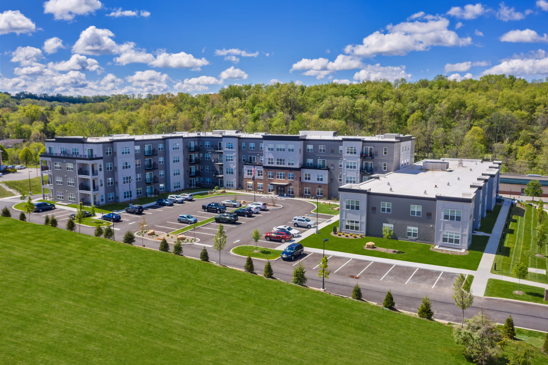 Aerial view of Ridgecrest of McMurray, a modern multi-story senior living facility surrounded by green lawns and trees under a blue sky with scattered clouds. The building has a large parking lot with several cars parked, and the facility is situated near a wooded area.