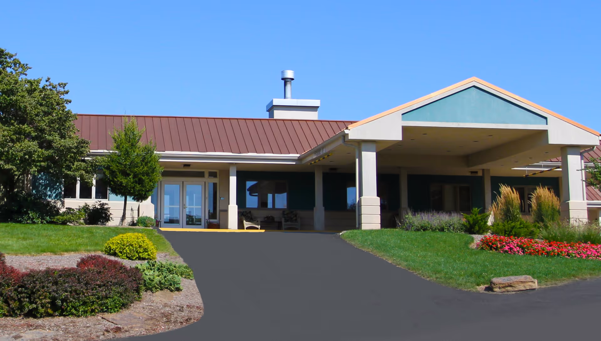 Front exterior view of Overlook Green Senior Living facility with a covered entrance, landscaped garden beds with shrubs and flowers, and a clear blue sky.