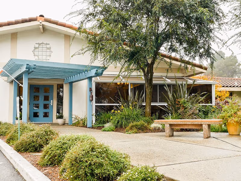 Exterior view of a single-story building with a blue entrance door under a pergola, surrounded by greenery, bushes, a tree, and a stone bench on a concrete walkway.