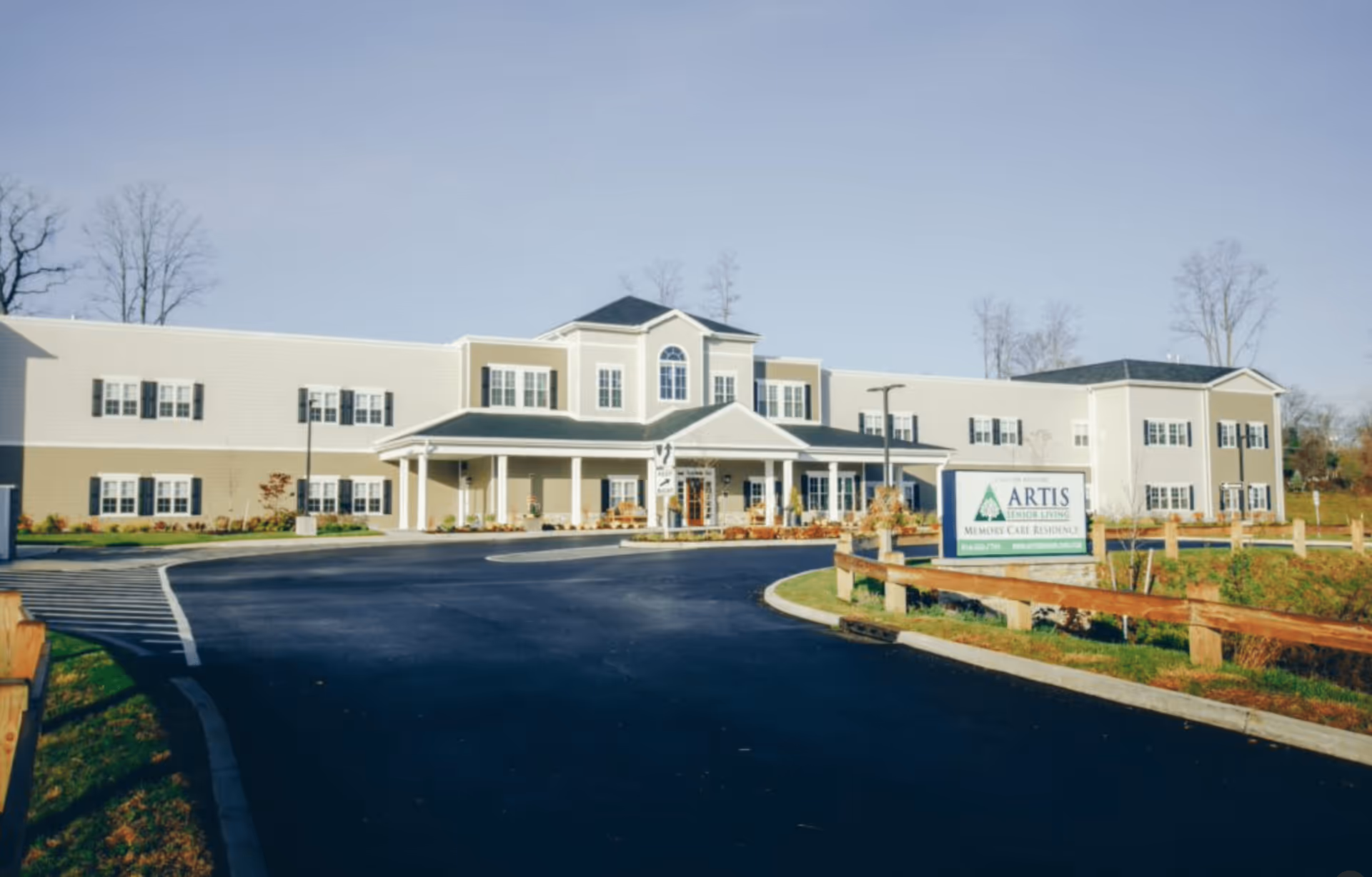 Exterior view of Artis Senior Living of Somers, a two-story beige building with white trim and multiple windows, a covered entrance, and a curved driveway leading up to the front. There is a sign in front indicating it is a memory care residence.
