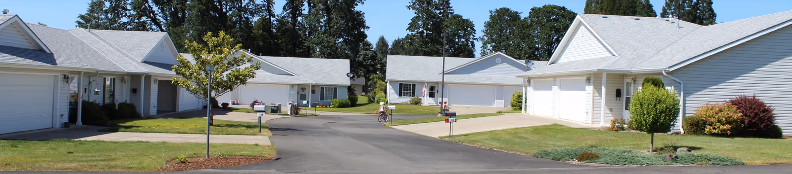 A quiet residential street in Marian Estates featuring single-story white houses with attached garages, well-maintained lawns, small trees, and a person riding a bicycle in the distance under a clear blue sky.
