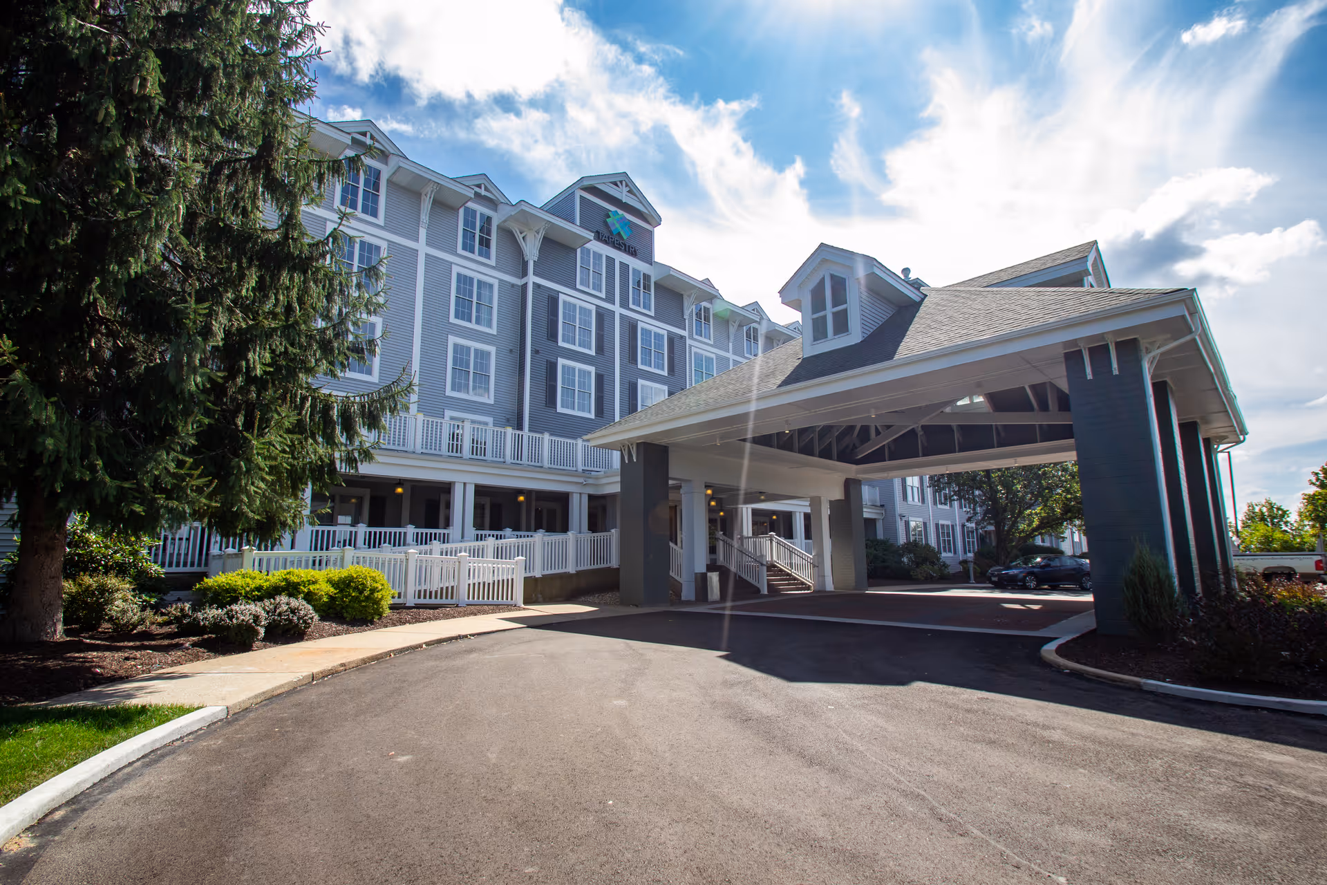 Exterior view of a senior living facility building with a covered entrance driveway, surrounded by landscaping and trees under a partly cloudy sky.