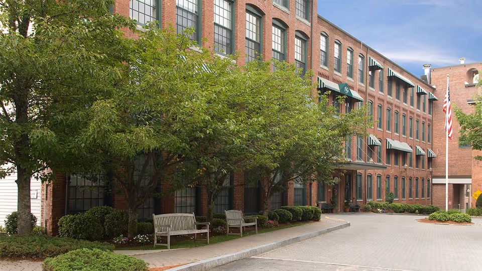 Exterior view of a multi-story brick building with large windows and green awnings. There are trees and bushes along the sidewalk, with two wooden benches placed under the trees. An American flag is visible on a flagpole near the building entrance.