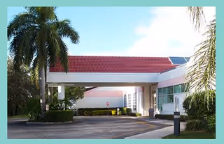 Exterior view of a building entrance with a red-tiled roof canopy, surrounded by palm trees and greenery under a partly cloudy sky.