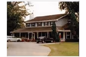 Exterior view of a two-story residential building with a covered porch, several cars parked in front, and trees surrounding the property.