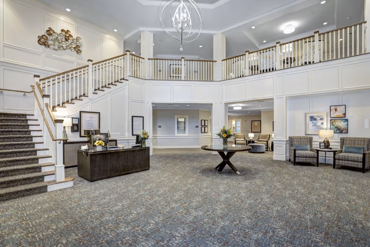 Spacious and elegant senior living facility lobby with a large carpeted floor, a dark wooden reception desk with two chairs, a round table with a flower vase in the center, a staircase with wooden handrails leading to an upper balcony area, and several seating areas with armchairs and framed artwork on the walls.