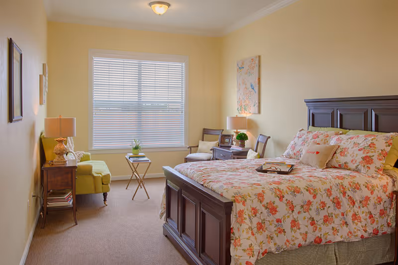 A cozy bedroom with a large wooden bed featuring floral bedding, two wooden chairs with cushions, a small wooden side table with a lamp and framed photo, a green armchair, a small folding table with a plant, and a window with closed blinds letting in natural light.