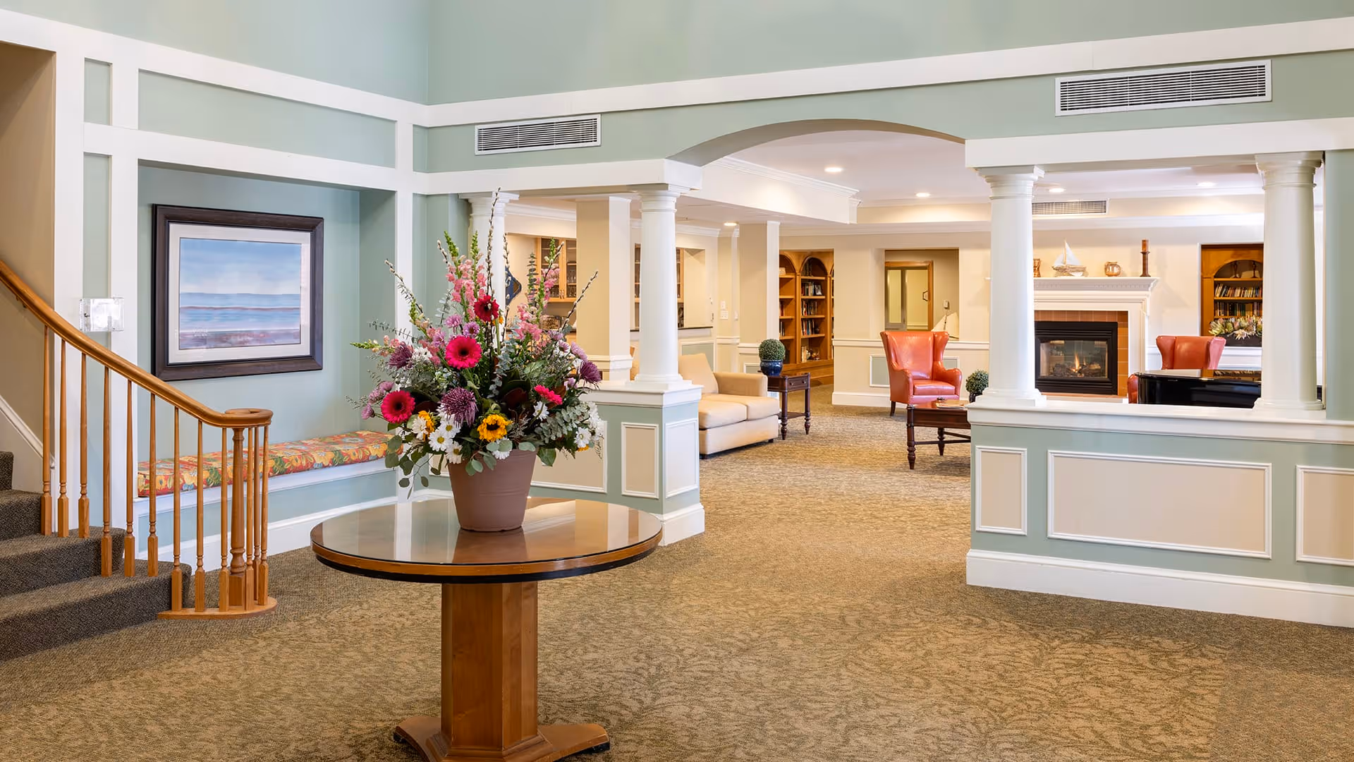 A spacious and well-lit senior living facility common area with a round wooden table holding a large floral arrangement in the foreground. To the left, there is a staircase with wooden railing and a cushioned bench beneath a framed painting. The background features a cozy seating area with beige sofas, red armchairs, a fireplace, and built-in bookshelves.