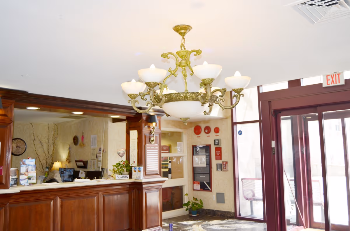 Interior view of a reception area with a wooden front desk, a decorative chandelier hanging from the ceiling, and large glass doors with an exit sign above them. The desk has various papers and signs, and there is a plant on the floor near the wall with fire safety equipment.