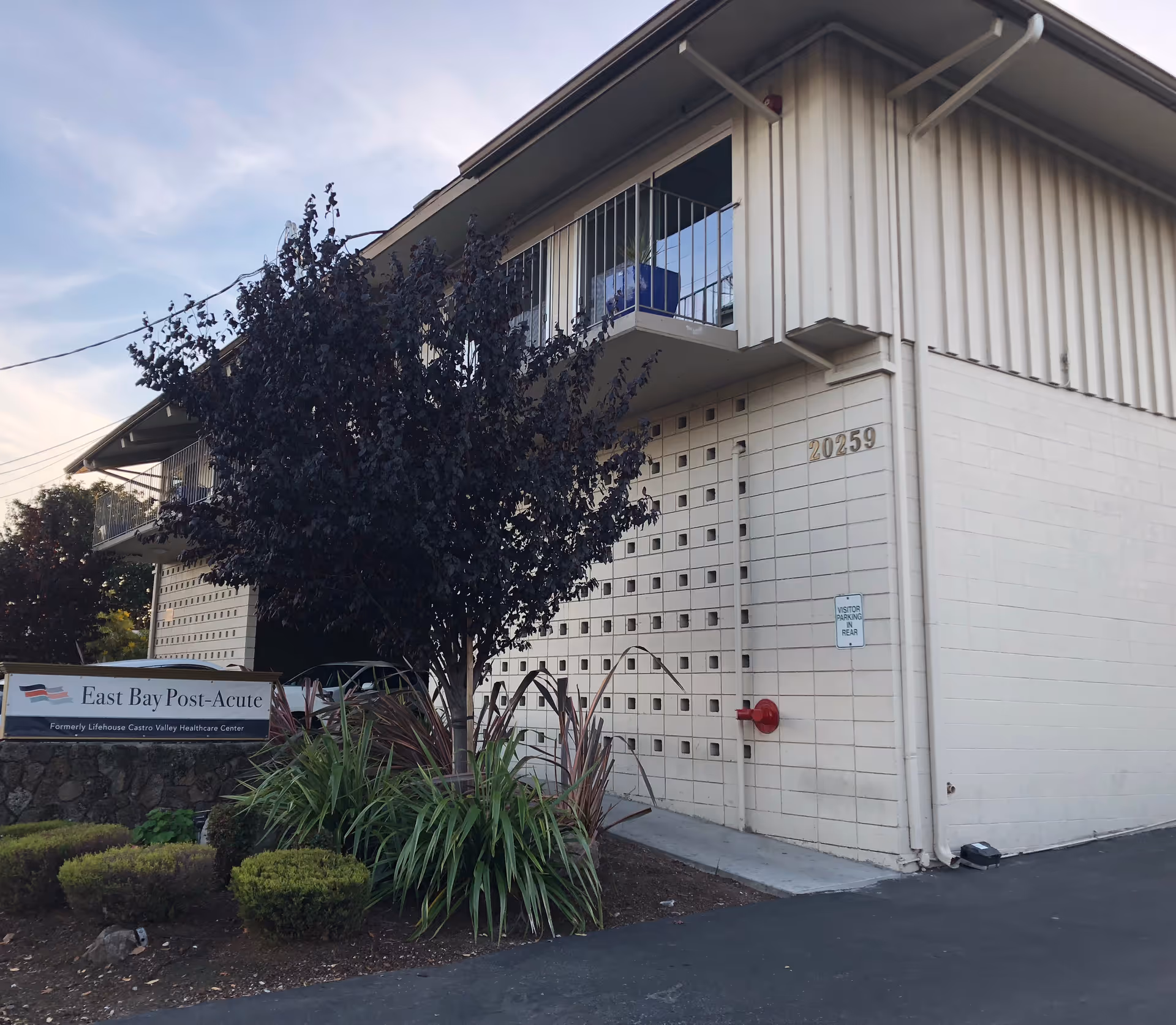 Exterior view of a two-story building with a balcony and a tree in front. A sign near the building reads 'East Bay Post-Acute, Formerly Lifehouse Castro Valley Healthcare Center.' The building number 20259 is visible on the wall.