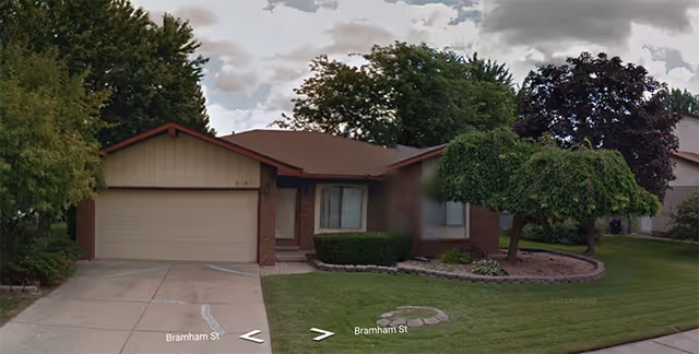 Single-story residential building with a two-car garage, a driveway, and a well-maintained front yard featuring trimmed bushes and trees under a cloudy sky.