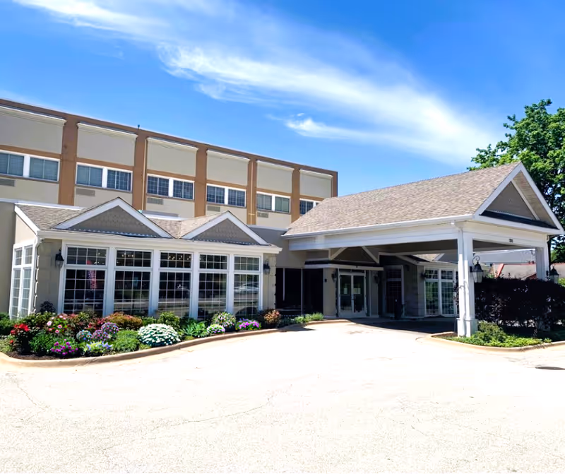 Exterior view of Plum Creek Supportive Living facility showing a multi-story building with large windows, a covered entrance, and a landscaped garden with colorful flowers under a bright blue sky.