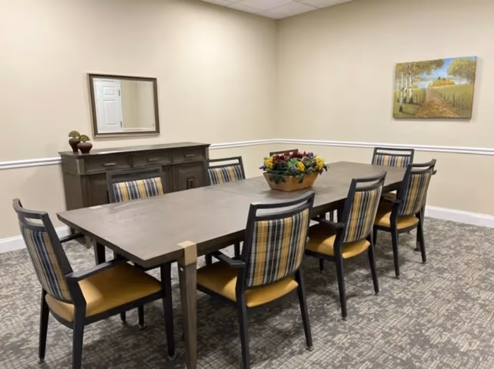 A dining room with a long rectangular wooden table surrounded by eight chairs with plaid backs and yellow seats. A decorative flower arrangement is centered on the table. Against the wall is a wooden sideboard with a mirror above it and two small potted plants. A landscape painting hangs on the adjacent wall. The room has beige walls with white trim and patterned carpet flooring.