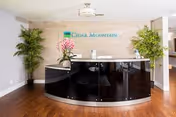 Reception area of Cedar Mountain Post Acute featuring a curved black reception desk with a computer and a pink orchid plant on top. The background has a light wood panel wall with the Cedar Mountain Post Acute logo mounted on it. There are two tall green plants on either side of the desk, and the floor is wooden.