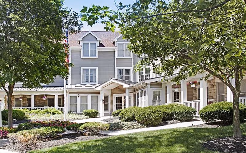 Exterior view of a senior living facility building with gray siding and white trim, surrounded by green trees, bushes, and a well-maintained garden. The entrance has a covered porch with white columns and a pathway leading to it.