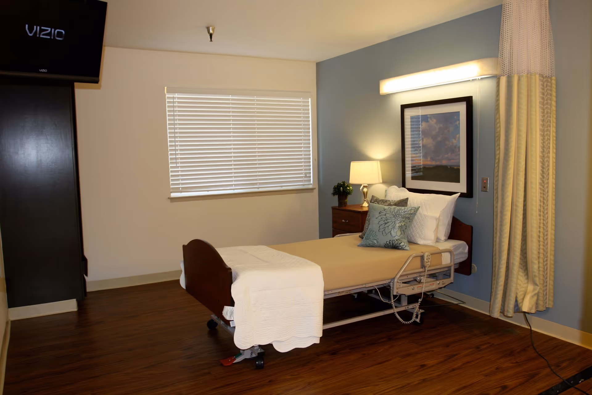 A healthcare facility bedroom with a single hospital bed made up with beige and white bedding and decorative pillows. There is a wooden nightstand with a lamp and a small plant next to the bed. A framed picture hangs on the blue accent wall behind the bed. A window with closed blinds is on the adjacent wall, and a flat-screen Vizio TV is mounted on the opposite wall. The floor is wood, and a privacy curtain is partially visible on the right side.