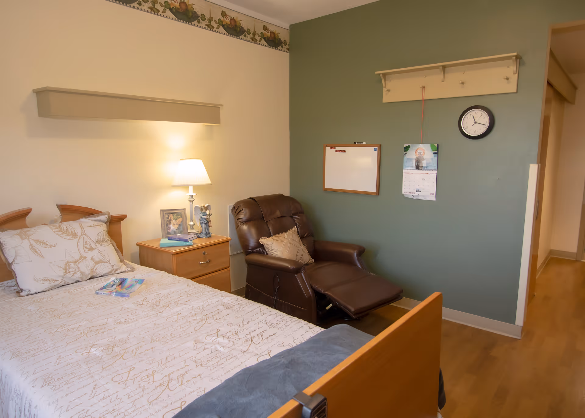 A cozy bedroom in a senior living facility featuring a single bed with patterned bedding, a wooden nightstand with a lamp, books, and a framed photo. Next to the nightstand is a brown recliner chair with a decorative pillow. The walls are painted in two tones, with a green accent wall displaying a clock, a calendar, a whiteboard, and a coat rack with hooks. The floor is wooden.