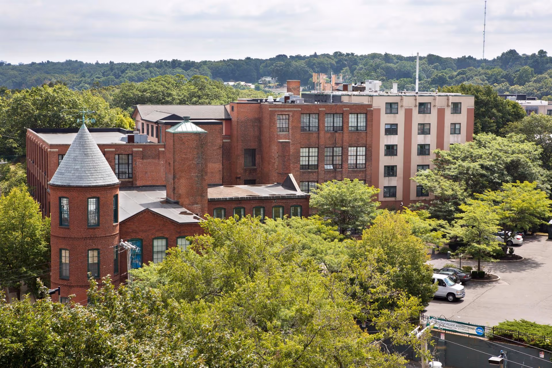 Exterior view of a multi-story brick building with a distinctive turret and several windows, surrounded by green trees and a parking lot with cars. The scene is set on a clear day with a wooded area in the background.