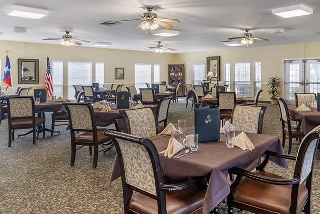 A spacious dining room with multiple tables covered in brown tablecloths, each set with folded napkins, glasses, and menus. The room has patterned carpet flooring, ceiling fans, and large windows allowing natural light. Flags and framed pictures decorate the walls.
