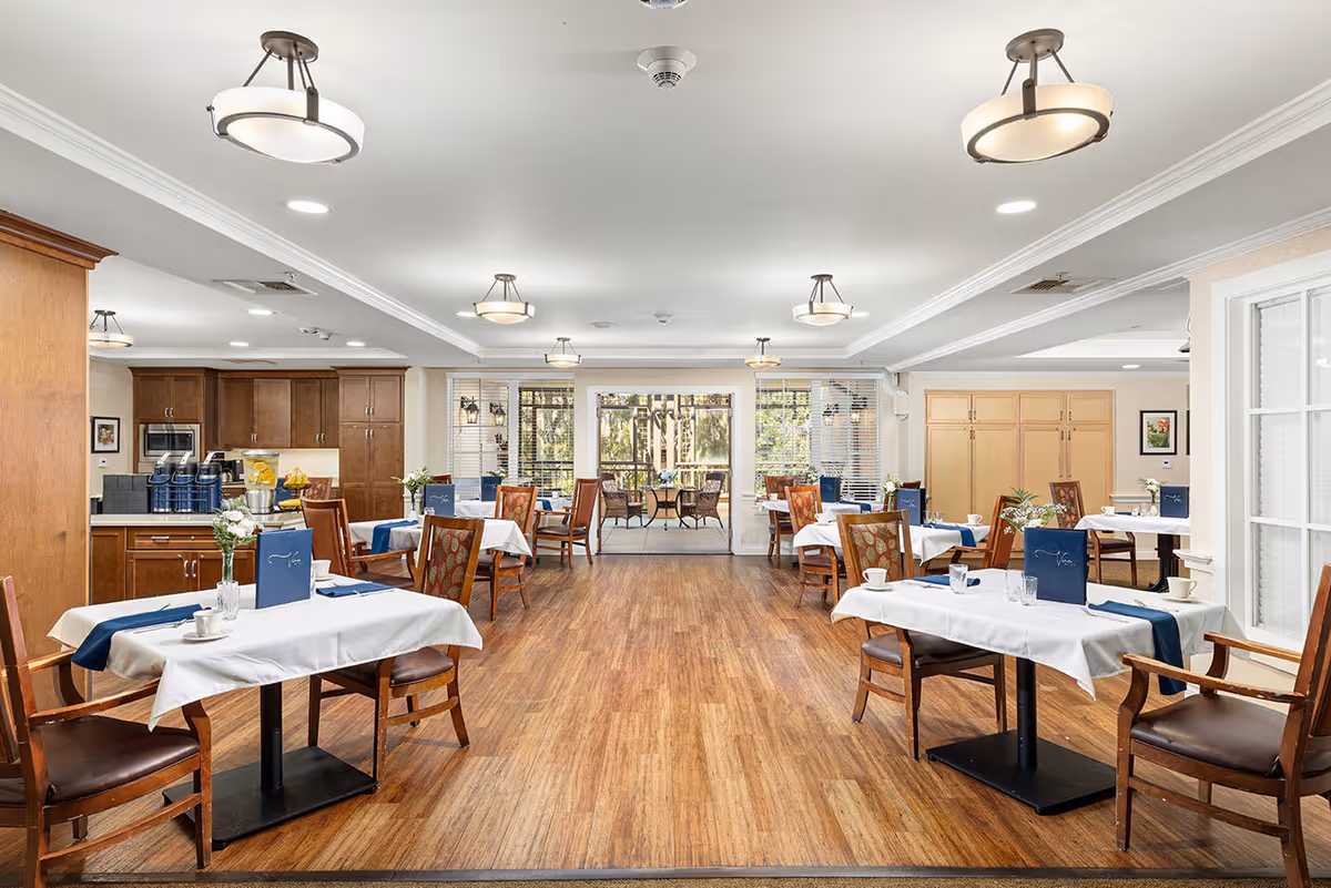 A bright and spacious dining room with wooden floors, multiple tables covered with white tablecloths and blue runners, wooden chairs with patterned cushions, and a kitchen area with wooden cabinets and coffee machines on the left side. Large windows and glass doors at the back provide a view of an outdoor patio with additional seating.