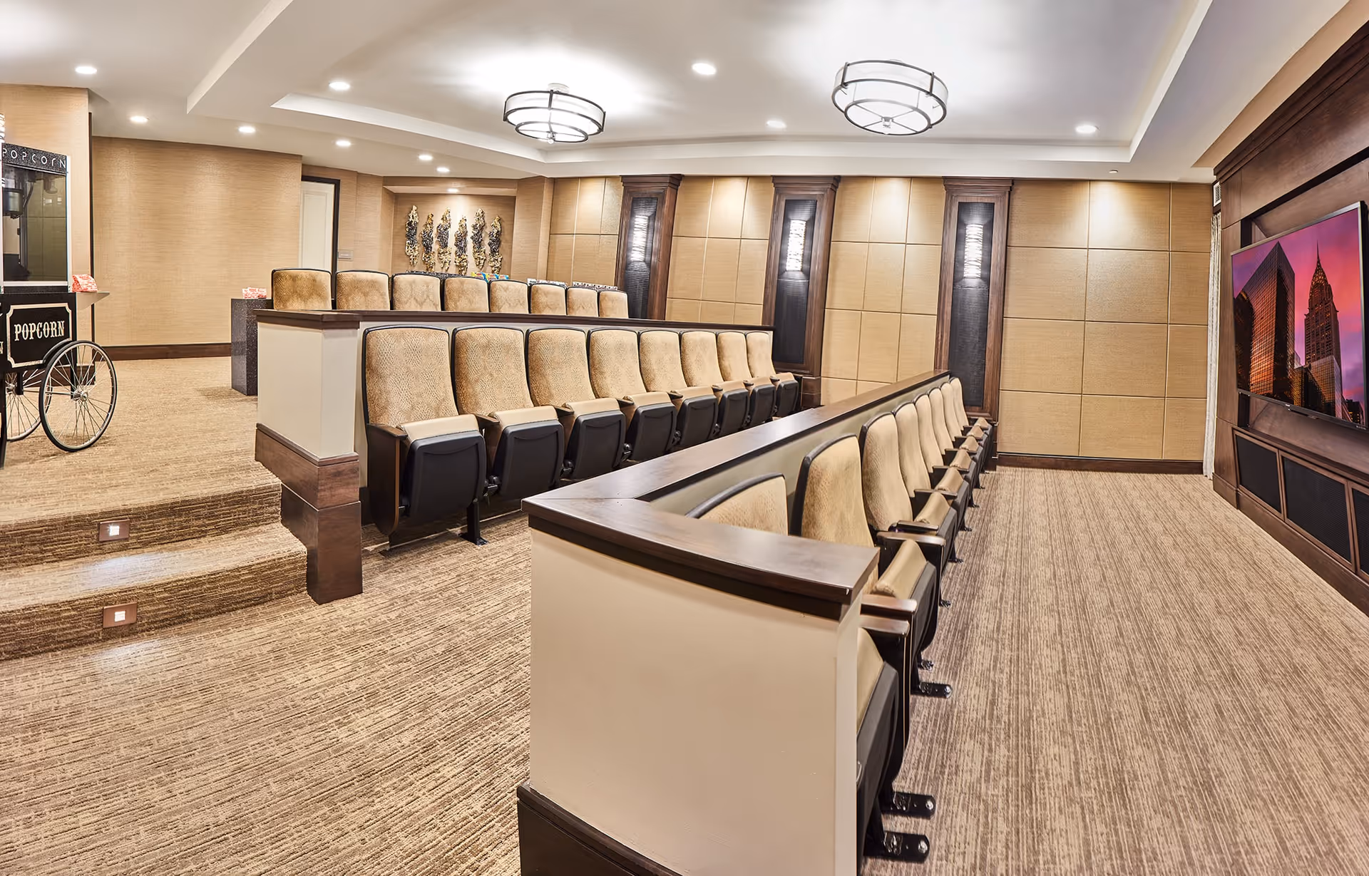 Interior view of a small theater room with tiered seating featuring beige cushioned chairs with dark wooden armrests. The room has beige walls with decorative panels and two round ceiling lights. A large flat-screen TV is mounted on the wall at the front, displaying an image of city buildings at sunset. A popcorn machine is visible on the left side near the entrance.