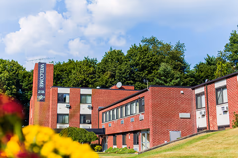 Exterior view of a red brick senior living facility building with multiple windows and air conditioning units. The building is surrounded by green grass, trees, and colorful flowers in the foreground under a partly cloudy blue sky. A vertical banner on the building reads 'WELCOME'.