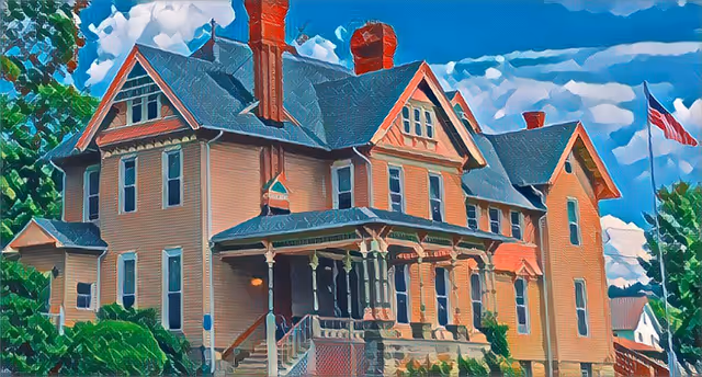 A large, multi-story historic building with beige siding, multiple chimneys, and a covered front porch with columns. The building is surrounded by greenery and trees, with an American flag flying on a flagpole to the right. The sky is blue with scattered clouds.