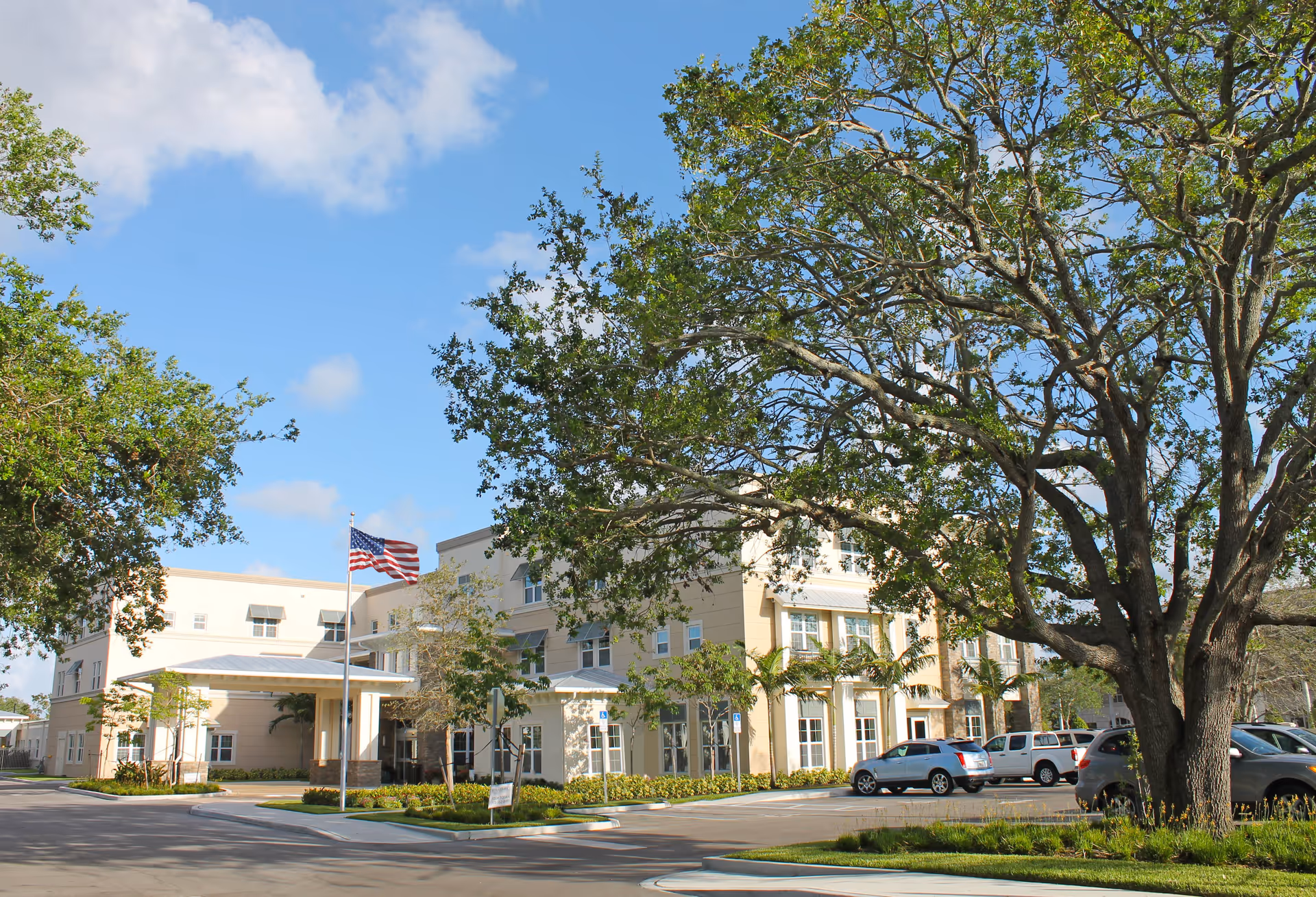 Exterior view of Renaissance Senior Living of Vero Beach, a multi-story beige building with white trim, surrounded by trees and a parking lot with several cars. An American flag is flying on a flagpole near the entrance under a covered driveway.