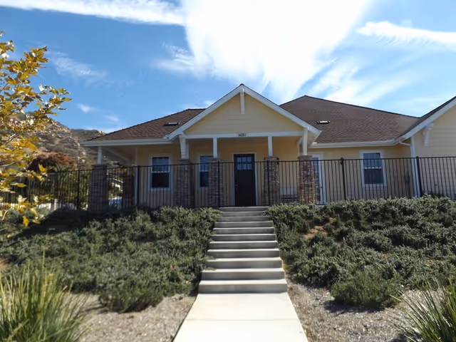 Front exterior view of a single-story yellow building with a brown roof, a black door, and several windows. There is a concrete staircase leading up to the entrance, flanked by green shrubbery and a black metal fence. The sky is blue with some wispy clouds.