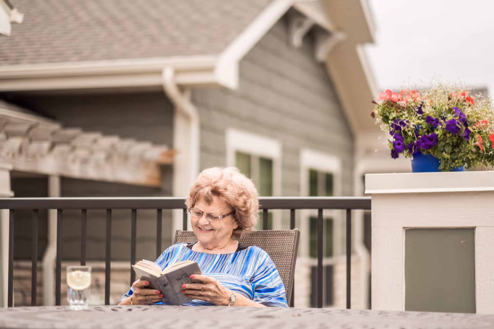 An elderly woman with glasses and curly hair is sitting outdoors on a patio chair, smiling while reading a book. There is a glass of water with a lemon slice on the table in front of her, and a planter with colorful flowers is visible on the right side. The background shows the exterior of a building with windows and a roof.