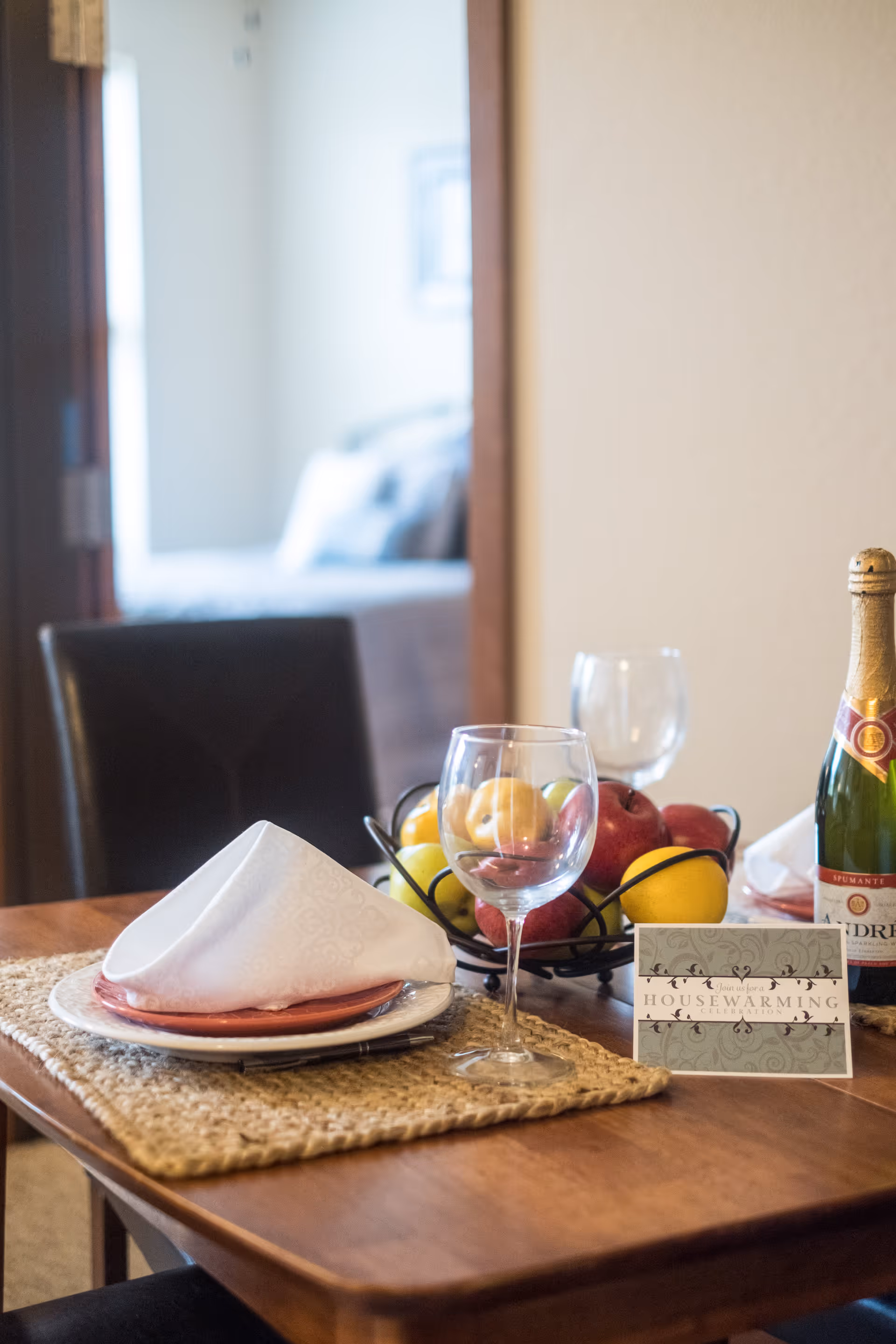 A dining table set with a woven placemat, a plate with a folded white napkin, a wine glass, a bowl of assorted fruits, a bottle of sparkling wine, and a housewarming card. In the background, a blurred view of a bedroom is visible through an open doorway.