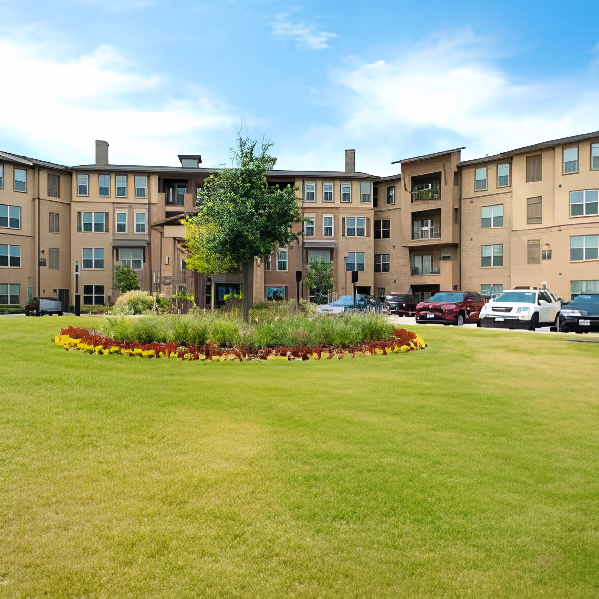 Exterior view of a multi-story senior living facility building with beige walls and numerous windows. In front of the building is a well-maintained lawn with a circular flower bed featuring green shrubs, red and yellow flowers, and a tree in the center. Several parked cars are visible on the right side near the building. The sky is partly cloudy with blue patches.