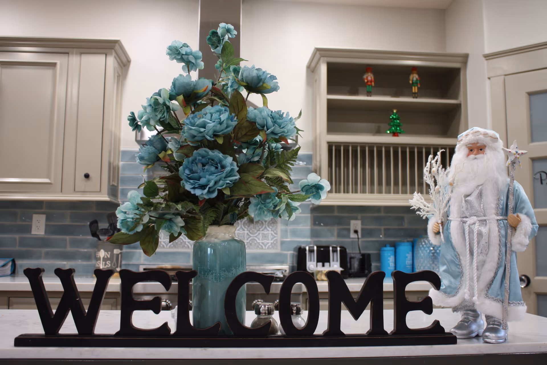 A kitchen countertop with a large vase of blue flowers and a decorative black 'WELCOME' sign. To the right of the sign is a figurine of Santa Claus dressed in a light blue and white outfit holding a staff. The background shows kitchen cabinets, a toaster, and some small decorative items including nutcracker figurines and a small Christmas tree.