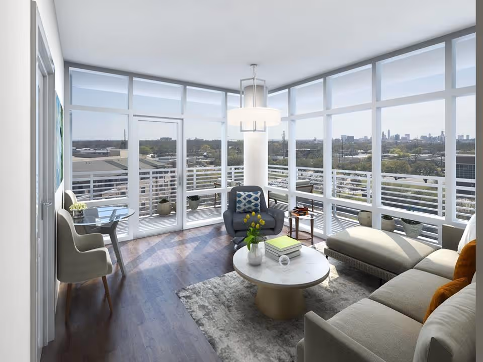 A bright living room with large floor-to-ceiling windows offering a panoramic city view. The room features a gray armchair with a patterned cushion, a round coffee table with books and a vase of yellow flowers, a sectional sofa with orange pillows, a small side table with books, and a small round table with two chairs near the window. The floor is wooden with a gray area rug, and a modern ceiling light fixture hangs above.