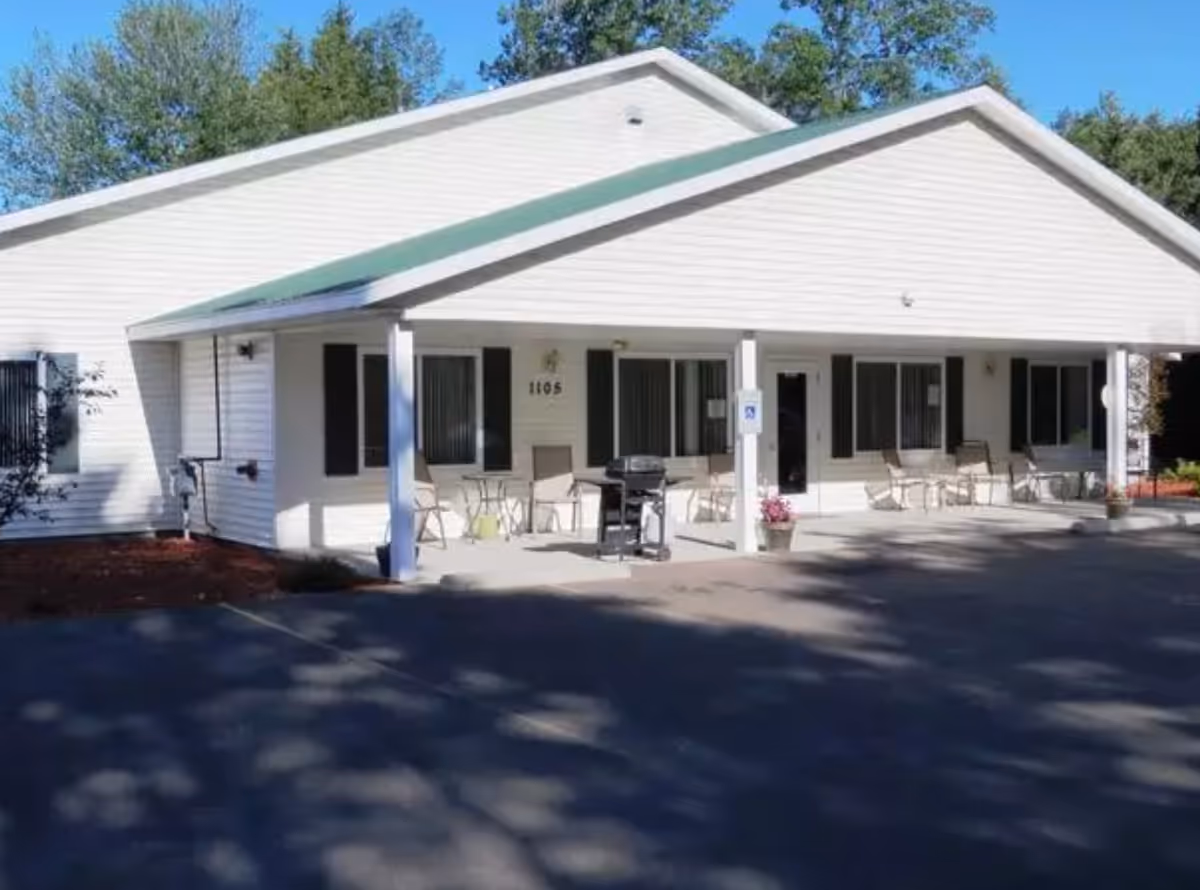 Exterior view of a single-story white building with a green roof and a covered porch area. Several chairs and small tables are arranged on the porch, along with a grill and potted plants. Trees are visible in the background under a clear blue sky.