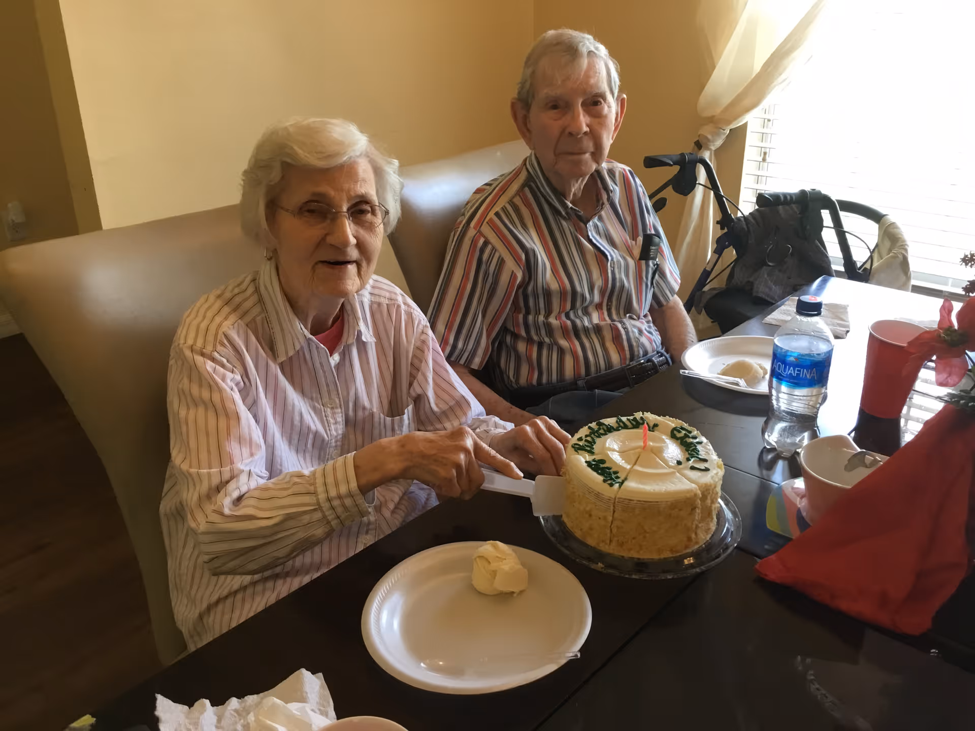 Two elderly individuals sitting at a dining table in a well-lit room. One woman is cutting a round cake with a single candle on it, while the man sits beside her. The table has a water bottle, paper plates, cups, and a red napkin with a flower decoration.