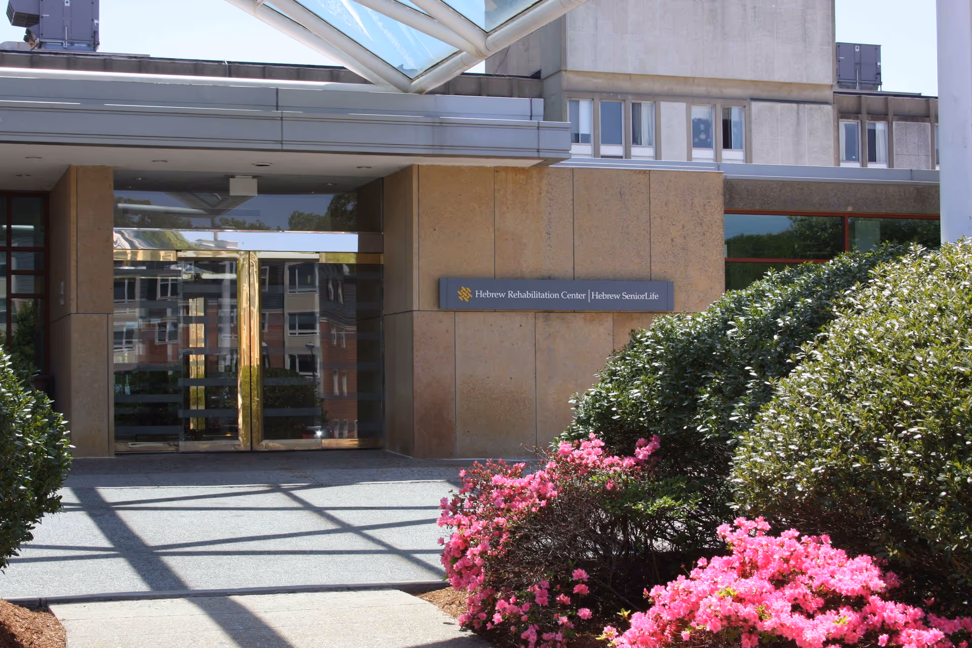Entrance to the Hebrew Rehabilitation Center in Boston with glass double doors, beige stone walls, and a sign displaying the facility's name. There are green bushes and pink flowers in the foreground.
