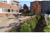 Outdoor patio area with several tables and chairs on a paved surface next to a brick building. There are plants and shrubs along the edge of the patio, and taller buildings are visible in the background under a clear sky.