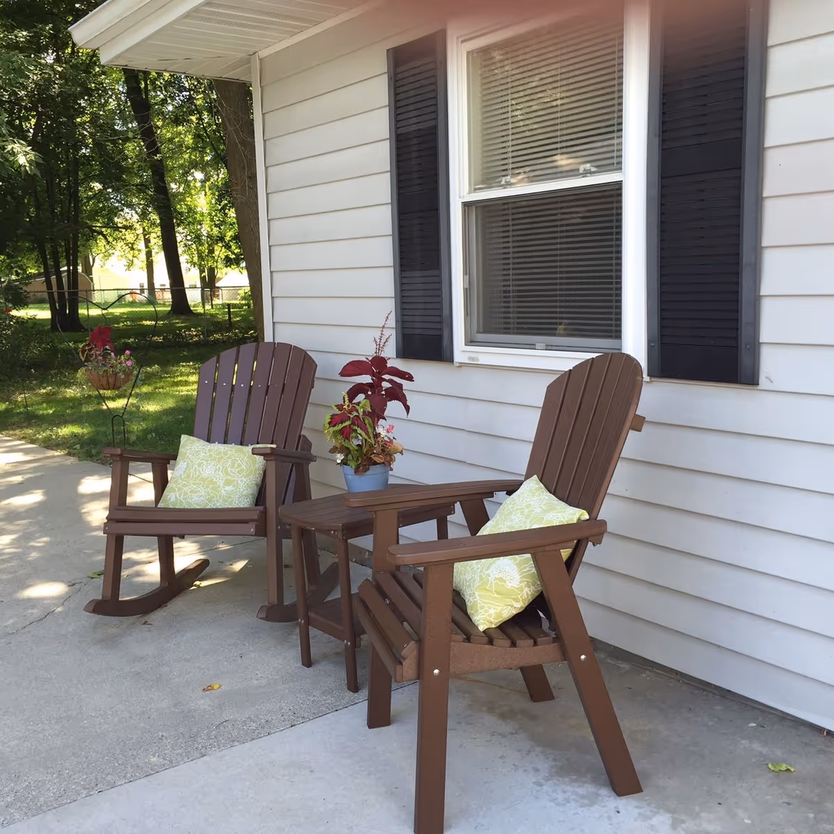 Two brown wooden chairs with light green patterned cushions are placed on a concrete patio outside a white house with black shutters. A small wooden table with a potted plant is between the chairs. Trees and greenery are visible in the background.