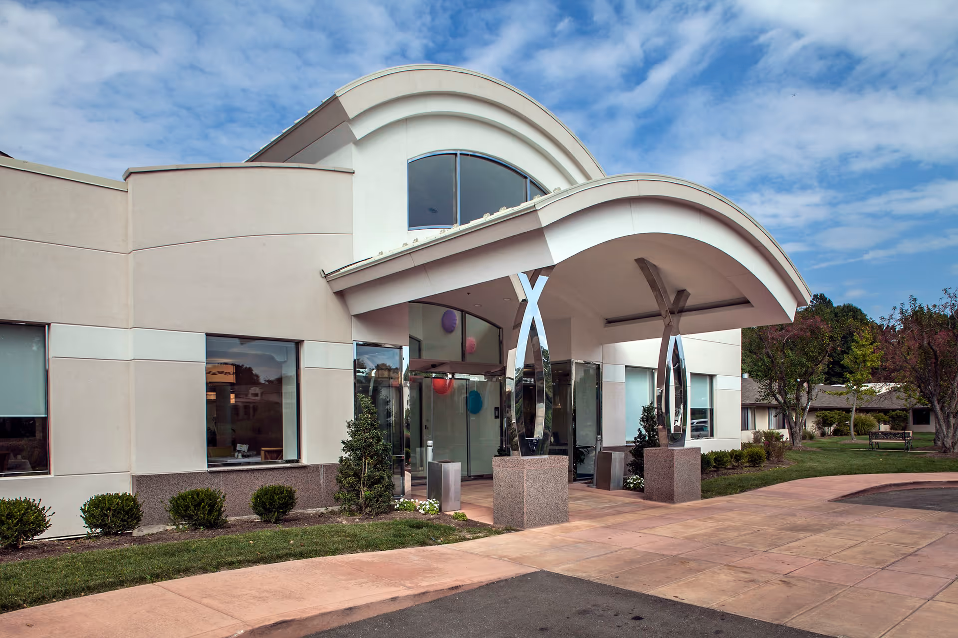 Entrance of a modern building with a curved white canopy supported by shiny metal pillars. The building has large windows and is surrounded by neatly trimmed bushes and a paved walkway. The sky is partly cloudy.