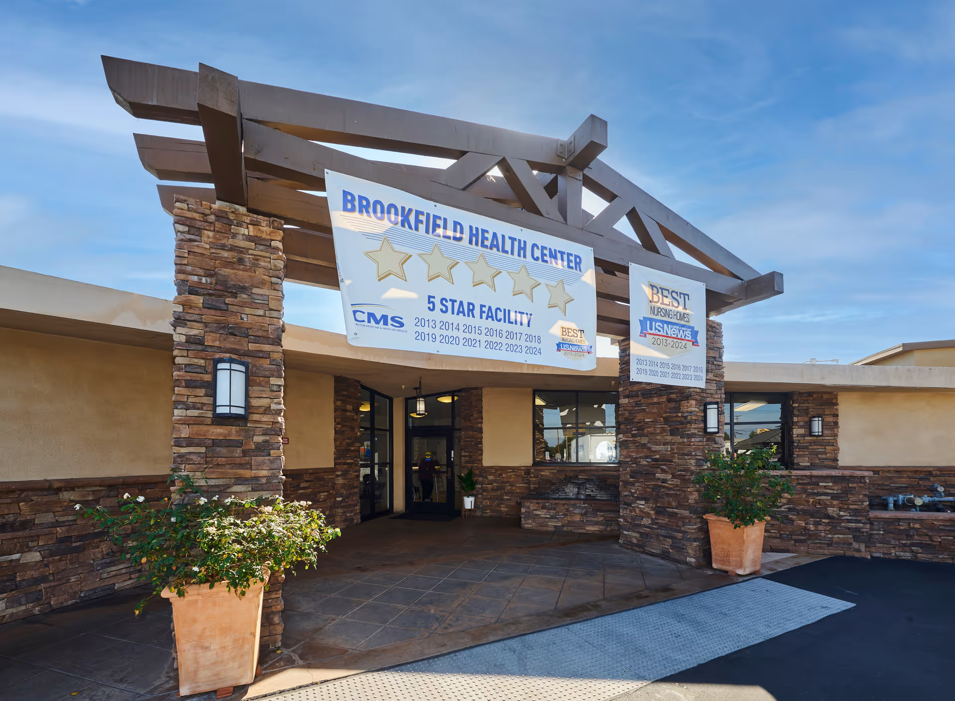 Entrance of Brookfield Health Center with stone pillars and a wooden pergola structure. Two large banners hang from the pergola, one displaying 'Brookfield Health Center 5 Star Facility' with CMS ratings from 2013 to 2024, and the other showing 'Best Nursing Homes US News 2013-2024'. There are potted plants on either side of the entrance and a clear blue sky above.
