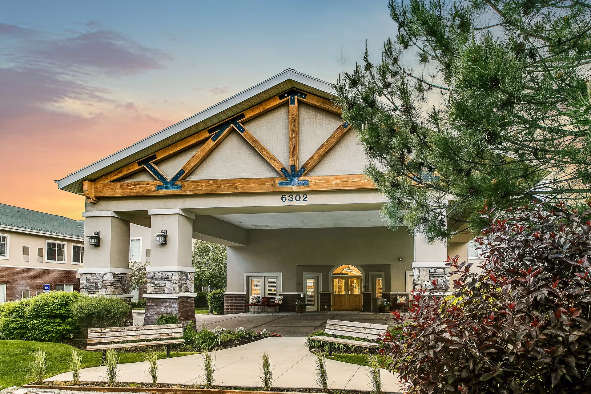 Entrance of a senior living facility with a covered driveway supported by stone and concrete pillars, wooden beams in the gable, benches on either side of the walkway, and landscaping with bushes and trees under a colorful sunset sky.