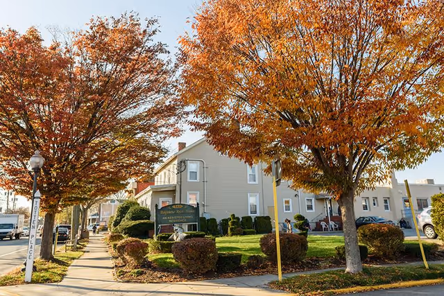Exterior view of Bayview Rest Home, an assisted living residence, showing a beige multi-story building with a well-maintained lawn and trimmed bushes. Trees with autumn-colored leaves line the sidewalk and street nearby.