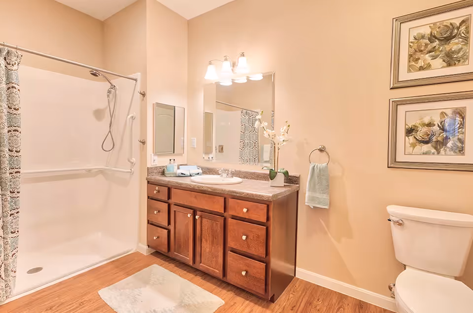 A clean and well-lit bathroom featuring a walk-in shower with a patterned shower curtain, a wooden vanity with a granite countertop and a sink, a large mirror with three light fixtures above it, a white toilet, two framed floral paintings on the wall, and a towel hanging on a ring.
