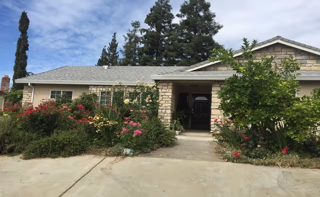 Front exterior view of a single-story building with a stone facade and a gray shingled roof, surrounded by lush green bushes and colorful flowering plants. Tall trees are visible in the background under a partly cloudy sky.