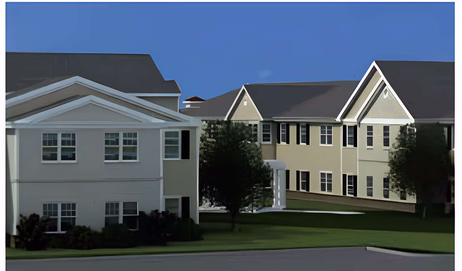 Exterior view of a senior living facility showing two connected buildings with multiple windows, pitched roofs, and a well-maintained lawn with trees in front under a clear blue sky.